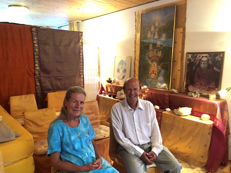 Eine Frau und ein Mann sitzen in einem Meditationsraum, an den Wänden hängen Bilder spiritueller Meister.